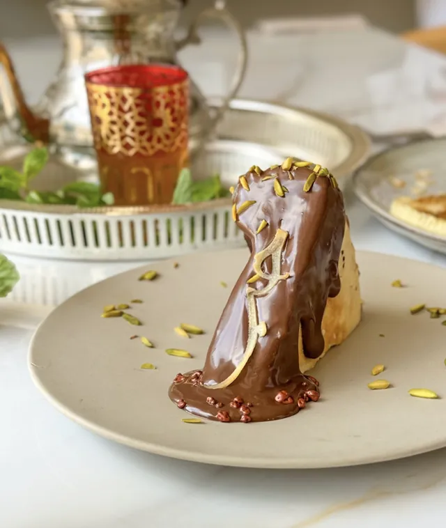 Dessert nappé de chocolat, pistaches et lettre en pâte d'amande, devant un service à thé